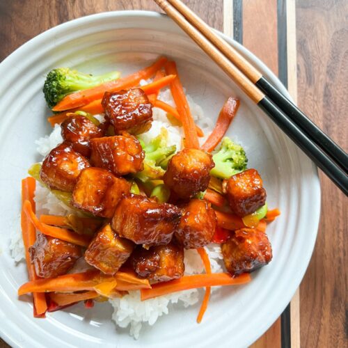 A white bowl contains a bed of rice topped with steam broccoli and carrots along with Crispy Marinated Tofu.