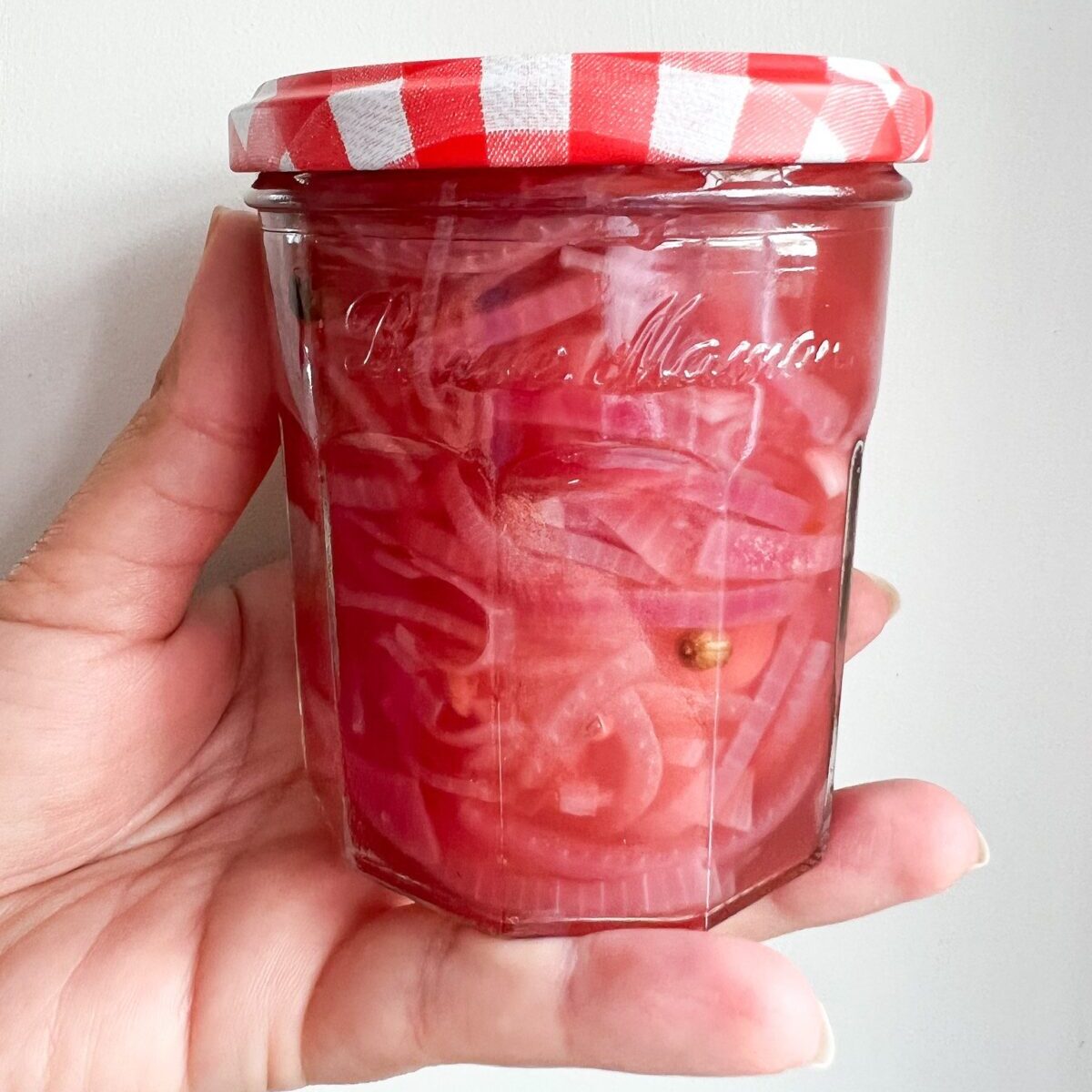 A woman's hand holds a jar of freshly made Pickled Red Onions. The hue is pink an peppercorns are seen within the simple brine.