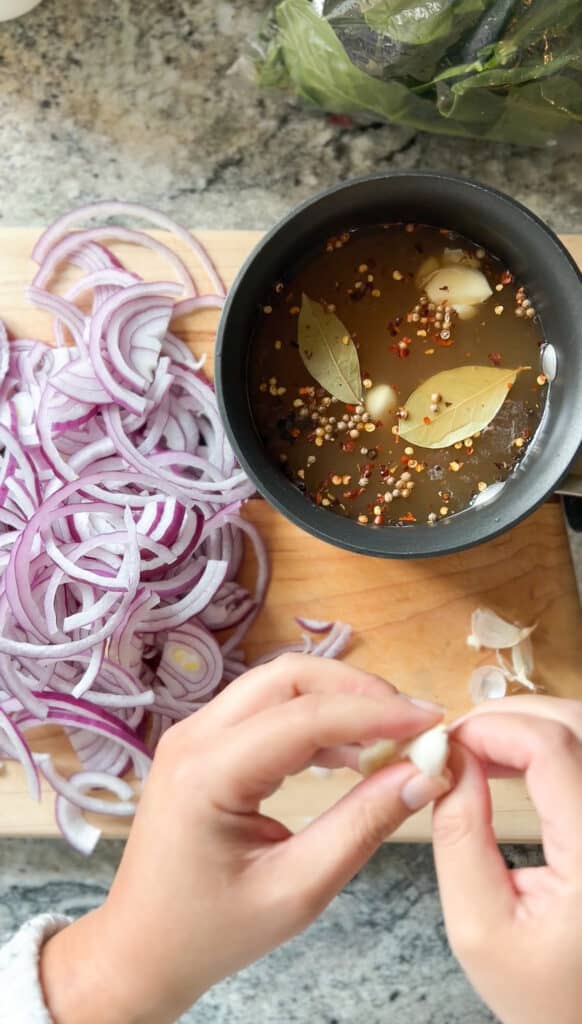 A pair of hands removes the outer skins from a clove of garlic in preparation for the brine for the pickled onions.