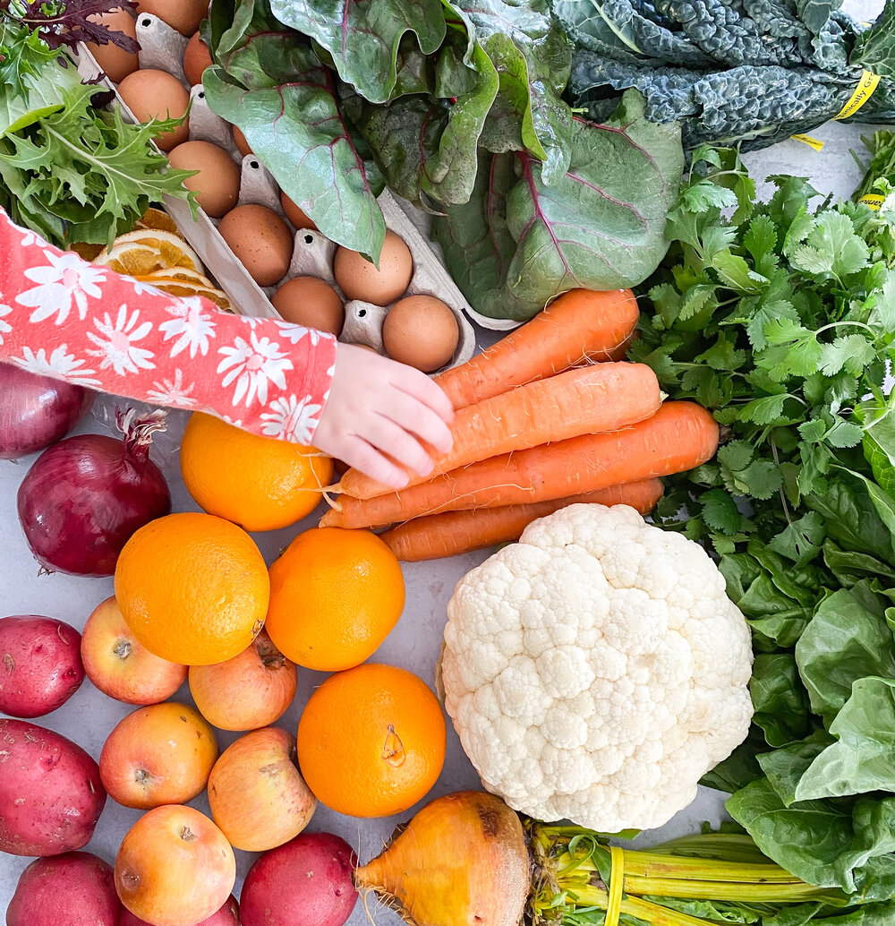 A toddler's hand reaches across the table of produce. Cauliflower, carrots, and citrus are visible.