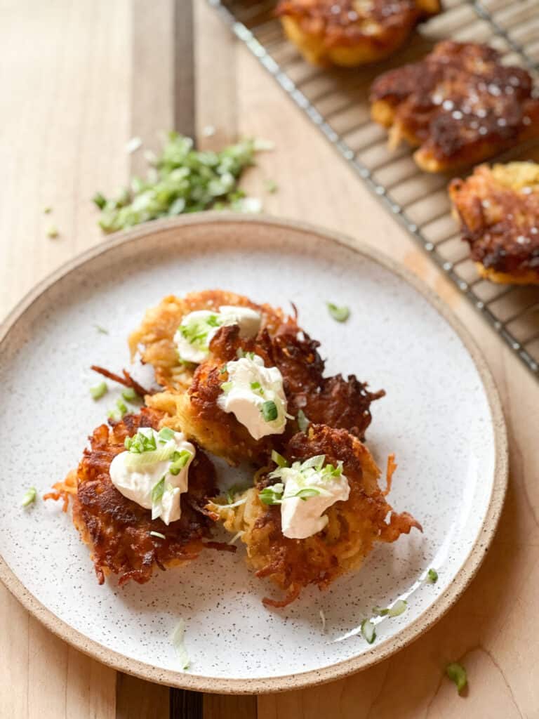 a white speckled plate holds 4 fried latkes garnished with sour cream and chopped scallions.
