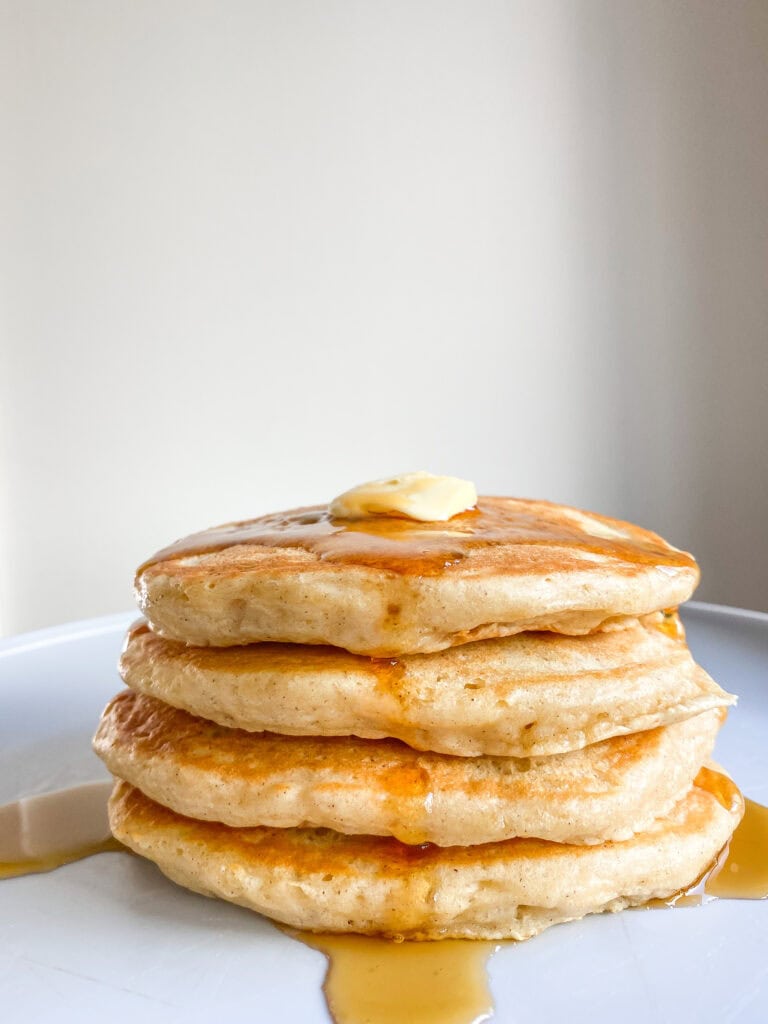 A close up shot of a short stack of 4 fluffy pancakes topped with a pad of butter and a drizzle of maple syrup. 