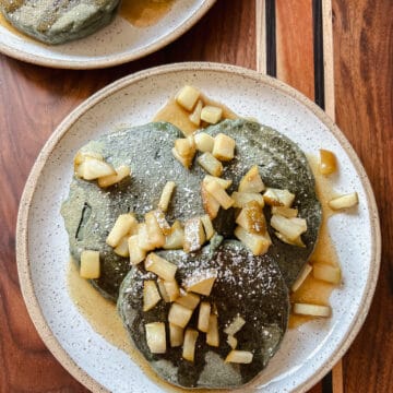 A speckled plate hold three blue corn pancakes drizzled with syrup and garnished with fresh seasonal chopped pears.