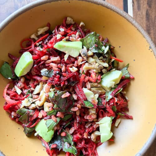 A bright orange bowl holds grated beet and carrot salad speckled with avocado and toasted walnuts.