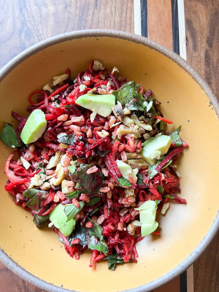 A bright orange bowl holds grated beet and carrot salad speckled with avocado and toasted walnuts.