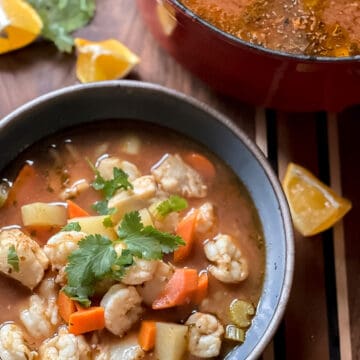 A bowl holds Mexican Fish Soup (Caldo de Pescado) garnished with limes and cilantro.