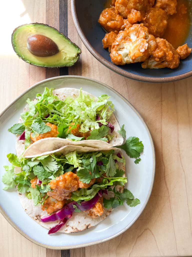 Direct overhead shot of a plate with two buffalo cauliflower tacos. Half an avocado is seen in the top left corner. 