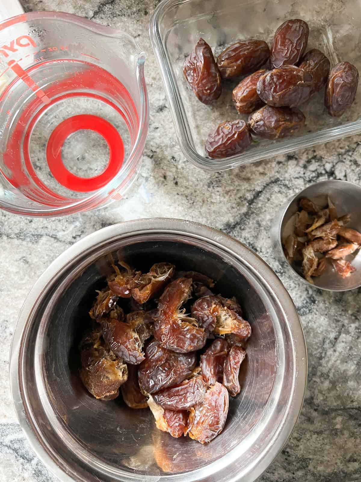 Whole dates and pitted dates in containers on a counter with a measuring glass of water to the side. 