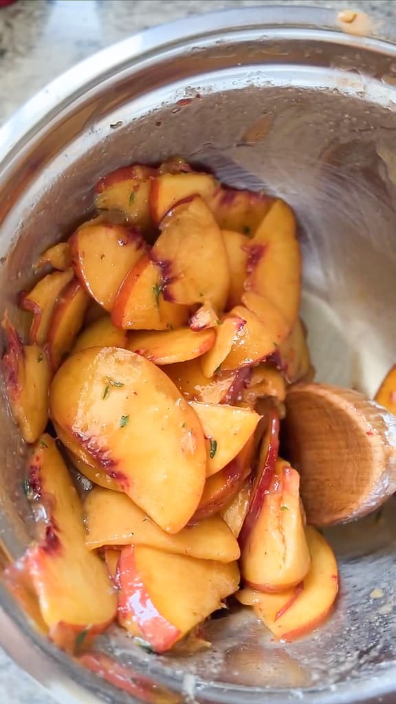 A large prep bowl holds sliced peaches seasoned in sugar and thyme. 