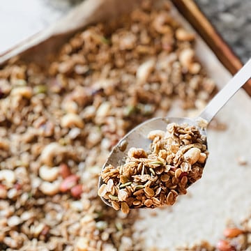 A batch of vegan maple cinnamon granola cools on a rimmed baking sheet with flecks of walnuts, almonds, cashews, and pumpkin seeds.