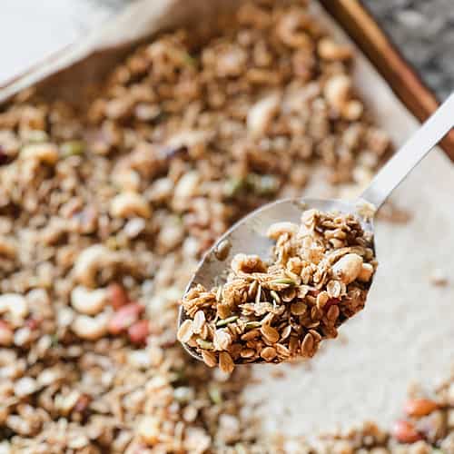 A batch of vegan maple cinnamon granola cools on a rimmed baking sheet with flecks of walnuts, almonds, cashews, and pumpkin seeds.