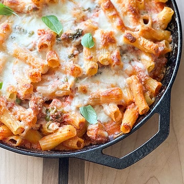A cast iron skillet contains a cheesy pasta bake with sausage and broccoli.