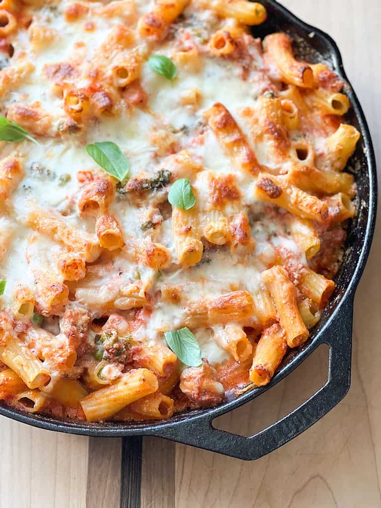 A cast iron skillet contains a cheesy pasta bake with sausage and broccoli.