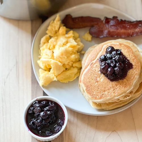 A plate of pancakes is garnished with blueberry compote with lemon and thyme served with a side of scrambled eggs and bacon.