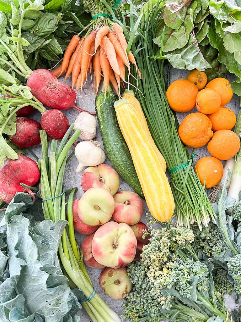 A table of produce is laid out containing donut peaches, zucchini, summertime citrus, radishes, and broccoli.