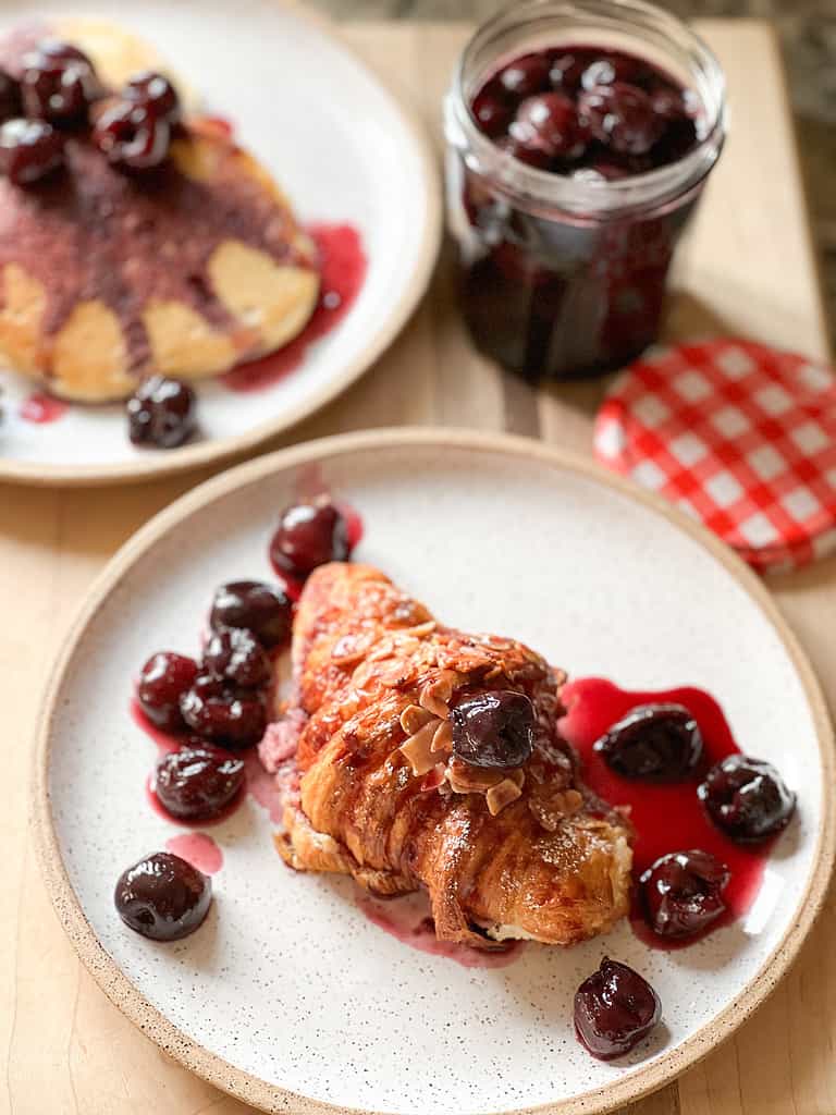 A white speckled plate holds an almond croissant topped with a homemade cherry compote.