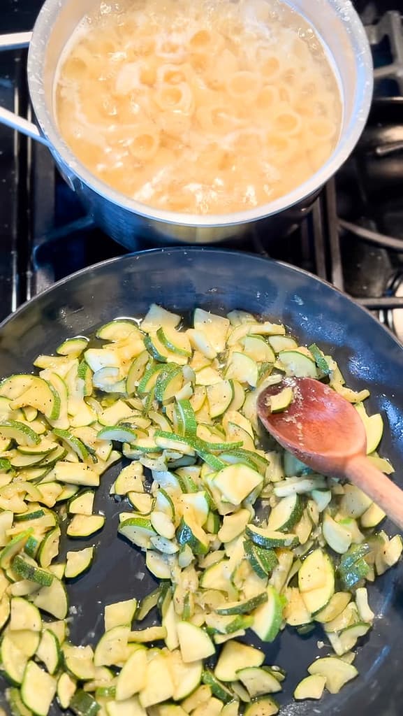 A nonstick skillet contains sliced zucchini in the process of caramelization. A sauce pan is above with pasta cooking until just al dente. 