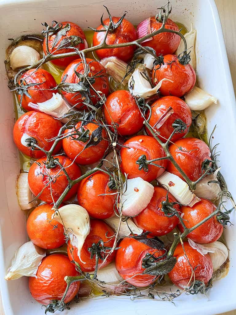 A white ceramic baking dish holds tomatoes, garlic, shallots, and herbs post roasting. 