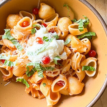 A large bowl is filled with pasta tossed in a roasted garlic tomato sauce garnished with parmesan, fresh tomatoes, and basil.