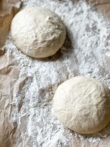 Two pizza dough balls sit atop a floured parchment lined baking sheet ready to proof for the weekly pizzas.