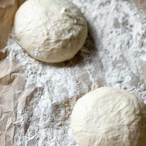 Two pizza dough balls sit atop a floured parchment lined baking sheet ready to proof for the weekly pizzas.