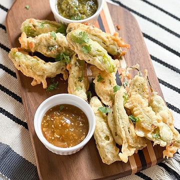 A wooden serving board displays a batch of shishito rellenos alongside two different types of salsas.