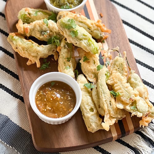 A wooden serving board displays a batch of shishito rellenos alongside two different types of salsas.