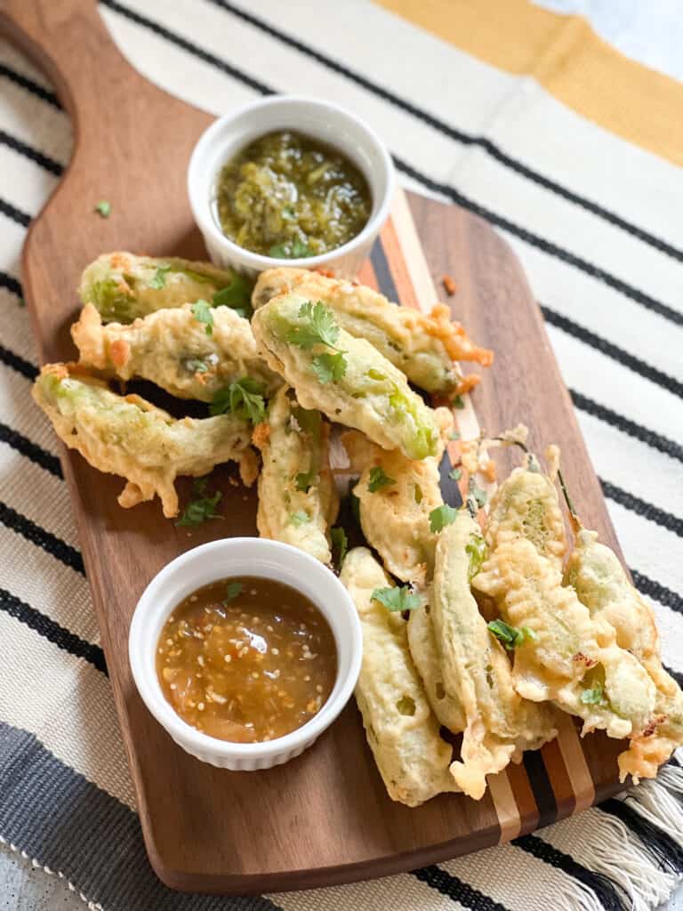 A wooden serving board displays a batch of shishito rellenos alongside two different types of salsas.