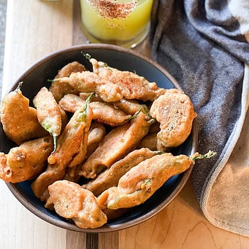 Portrait photo of a shallow bowl cradling a fresh batch of Shishito Rellenos. Two mezcal margaritas appear in the upper left corner to complete this happy hour snack.