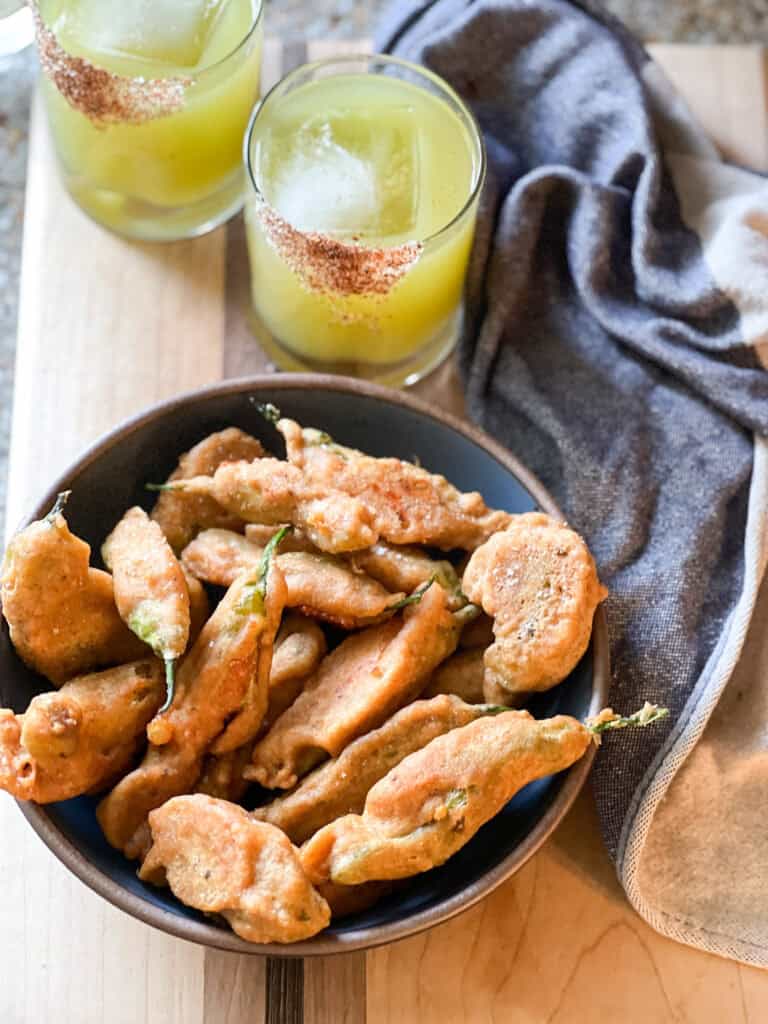 Portrait photo of a shallow bowl cradling a fresh batch of Shishito Rellenos. Two mezcal margaritas appear in the upper left corner to complete this happy hour snack.