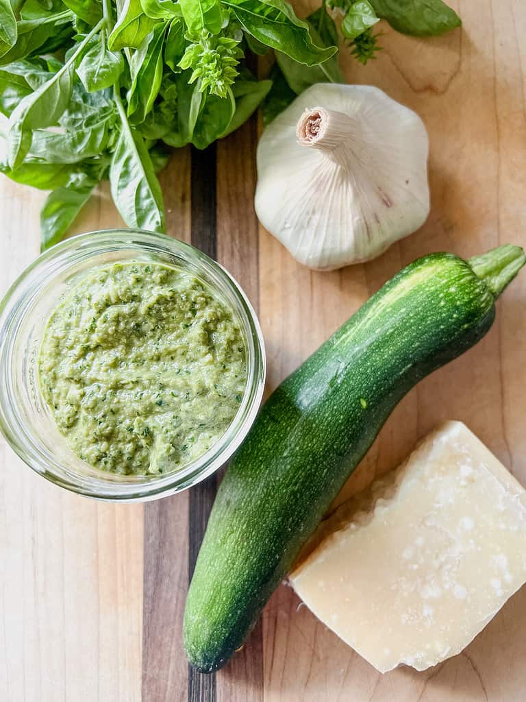 A clear jar of fresh made zucchini and basil pesto sits on top of a wooden cutting board accented with the ingredients involved to make this delicious sauce. 