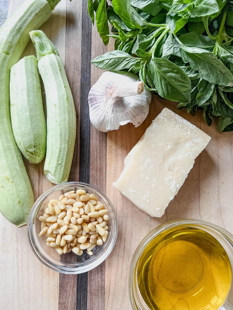 A wooden cutting board displays summertime zucchini alongside toasted pine nuts, parmesan, olive oil, garlic, and basil before transforming into pesto.