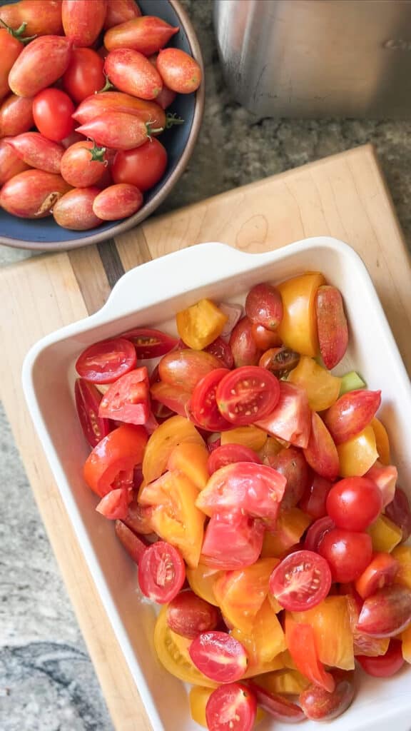 A white ceramic baking dish contains quartered and halved heirloom tomatoes in a mixture of yellows, oranges, and reds, in preparation for roasting. 