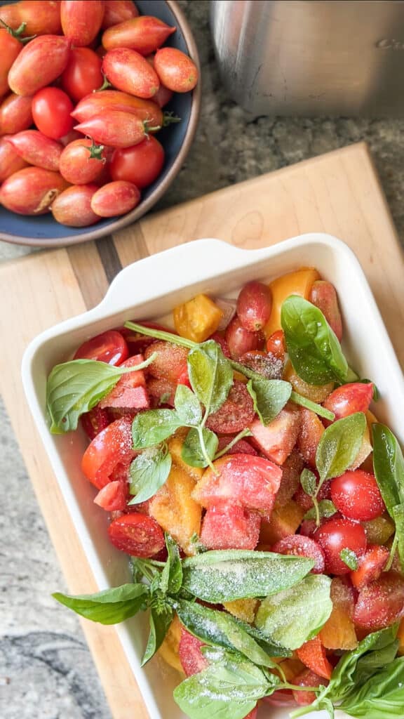 A white ceramic baking dish contains quartered and halved heirloom tomatoes in a mixture of yellows, oranges, and reds, in preparation for roasting. Fresh basil leaves are garnished on top prior to roasting. 