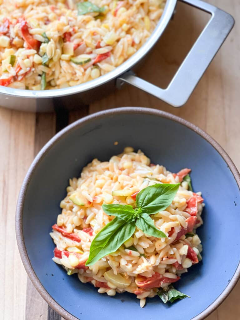 A bowl of one-pot orzo with tomatoes, corn, and zucchini is garnish with fresh basil and grated parmesan. 