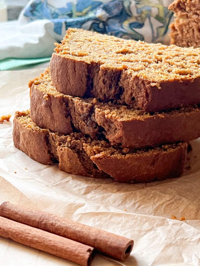 Freshly baked slices of one bowl pumpkin bread are stacked on top of one another gently. A pair of cinnamon sticks are laid in front of the stacked slices. 