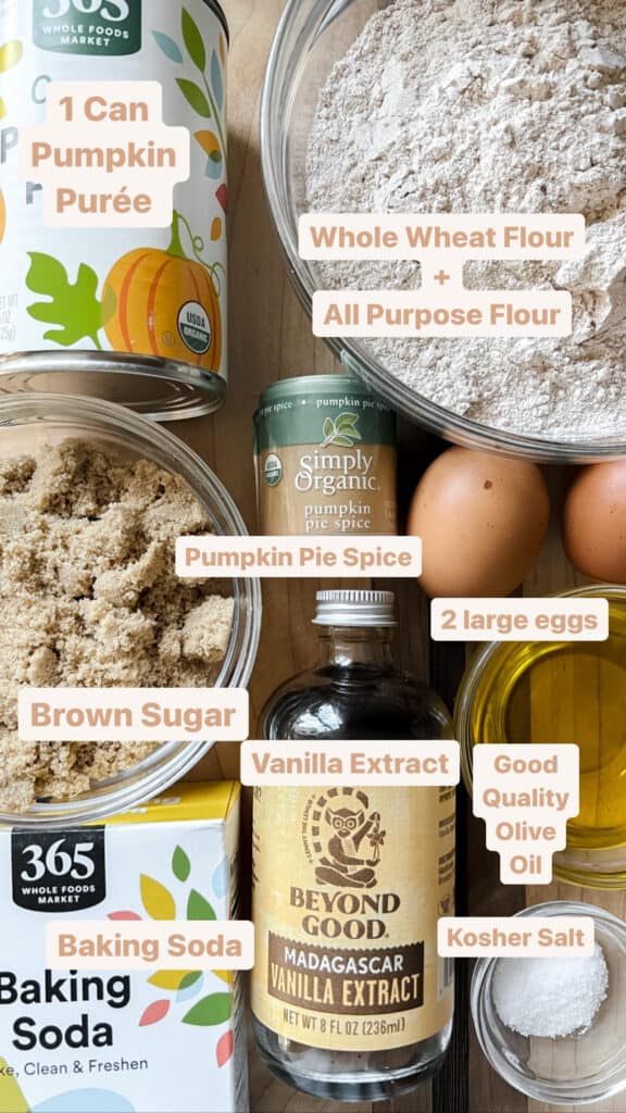 A flatlay image of all the ingredients featured in this one-bowl pumpkin bread. The labels read: 1 can pumpkin puree, whole wheat flour + all purpose flour, brown sugar, pumpkin pie spice, vanilla extract, good quality olive oil, kosher salt, and baking soda.
