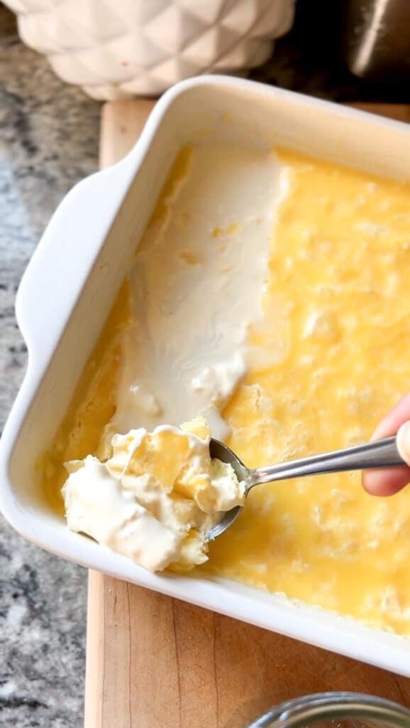 Phase 2 in the recipe for homemade clotted cream: allowing cream to set. A white ceramic dish  contains heavy cream that's been setting for 12 hours. A silver spoon is seen scooping up the separated milkfat that's now the clotted cream. 