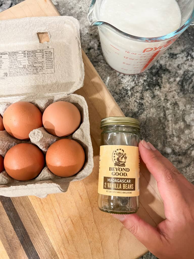 A flat lay image contains 4 brown eggs, a measuring cup with 2 cups of heavy cream. A hand holds steady a jar of vanilla beans with the label reading "Beyond Good: Madagascar 2 Vanilla Beans" to be used for the small batch vanilla bean ice cream. 