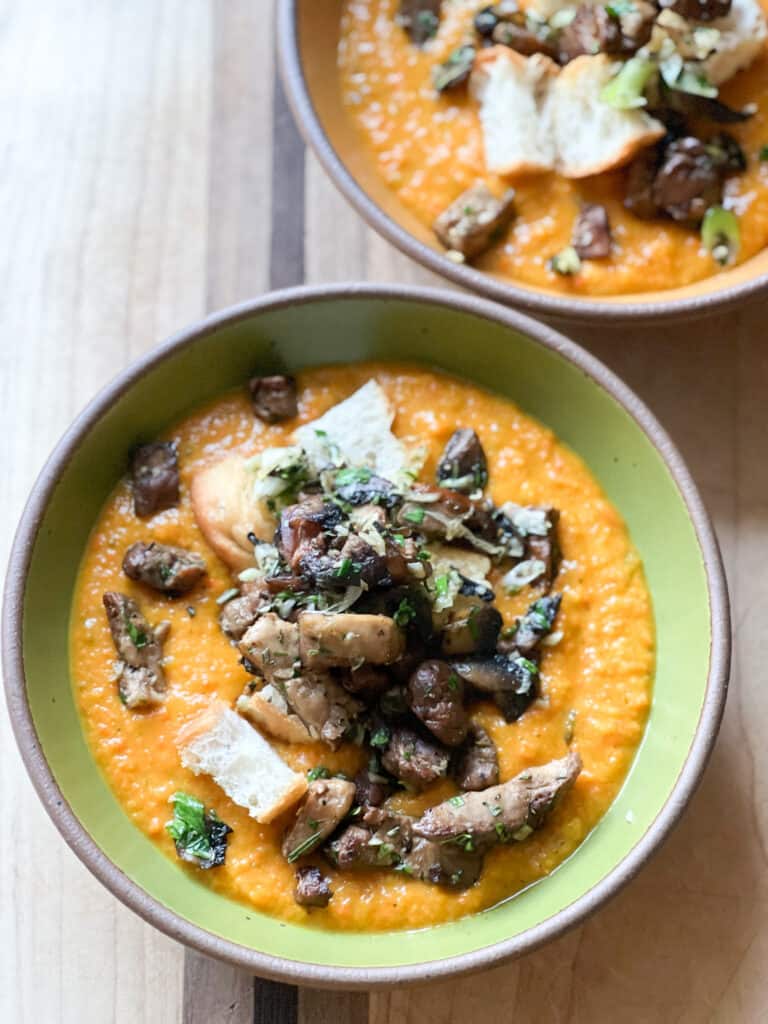 A vibrant green soup bowl holds Maple Harissa Roast Carrot soup topped with Crispy Mushrooms and Focaccia Croutons.