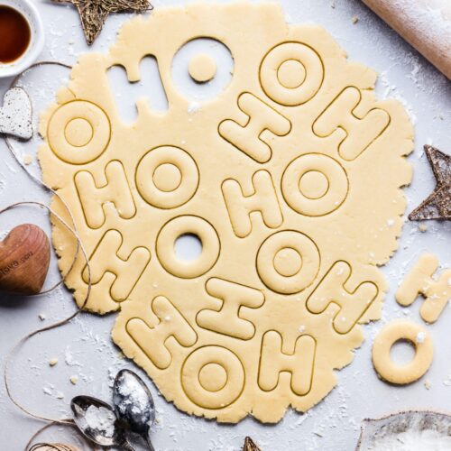 A flatlay image of pre-cut homemade sugar cookie dough. Shapes are the letters H and O for "ho, ho, ho."