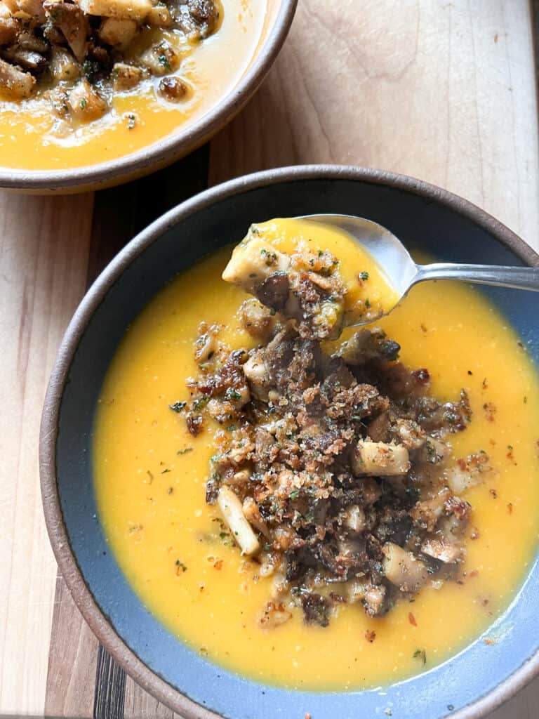 A spoon holds a spoonful of Spiced Squash Soup above a bowl filled with the soup and topped with Crispy Mushrooms. 