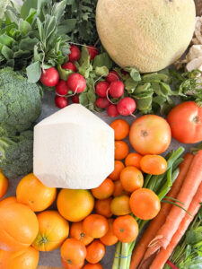 A flatlay image of seasonal produce featuring coconut, carrots, spring citrus, and fava bean greens.
