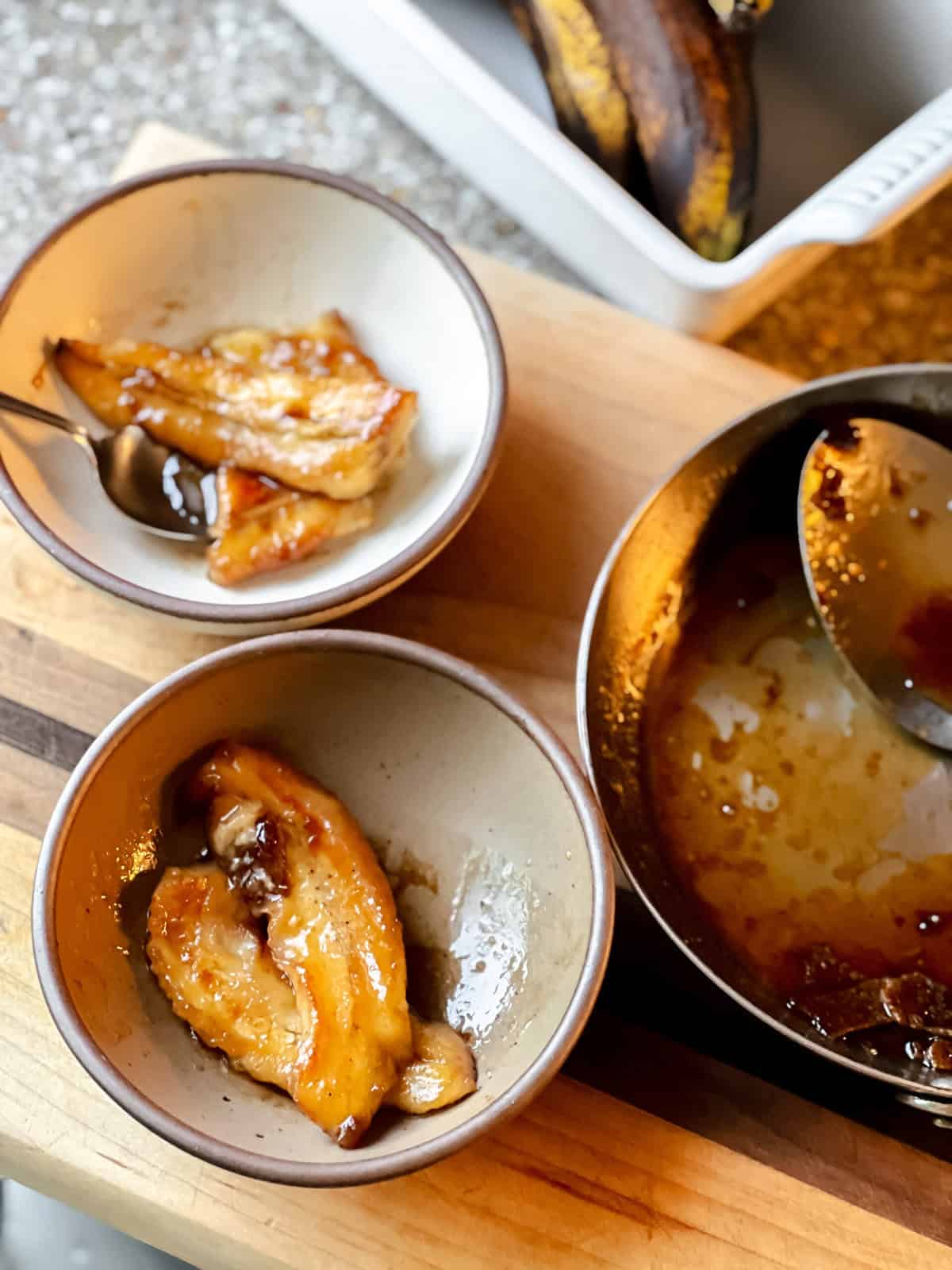 Banana fosters in bowls on a table next to a pan with some foster sauce still clinging to the sides.
