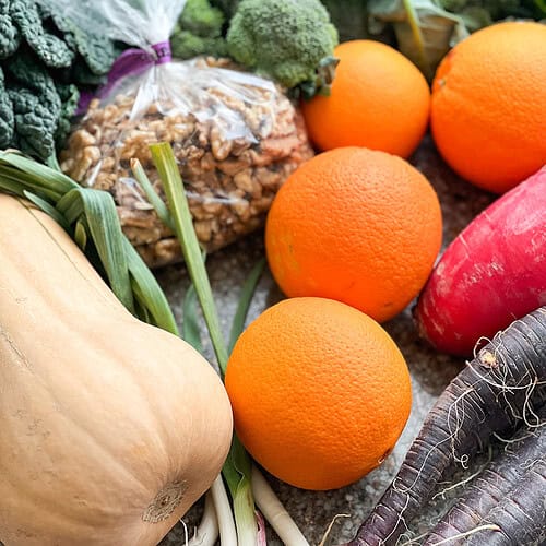 Portrait Photo of late winter produce for Dinner Plans Week 9 featuring red daikon radish, purple carrots, 4 navel oranges, a few small heads of broccoli, 1 butternut squash, a small bunch of tuscan kale, and a bag of fresh California walnuts.