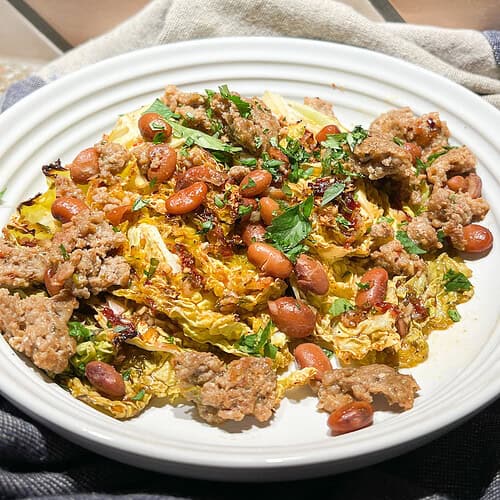 Portrait image of a shallow white pasta bowl filled with Roasted Baby Cabbage marinated in chipotle and lime. The cabbage is topped with cooked Mantequilla beans and Pork Chorizo.