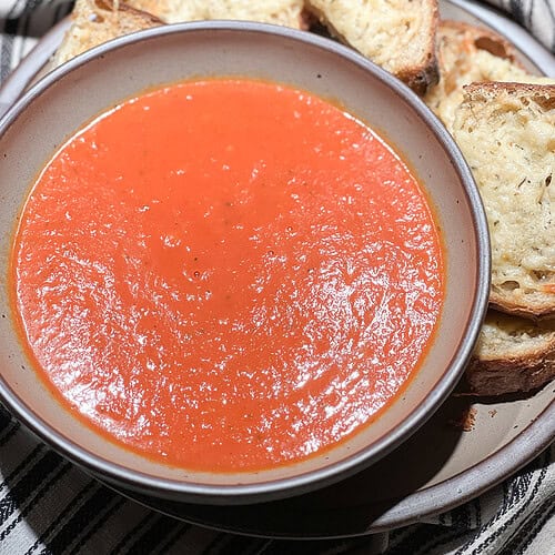 Portrait photo of Homemade Roast Tomato Soup filled in a soup bowl with a side of Gruyere and Parmesan toast.