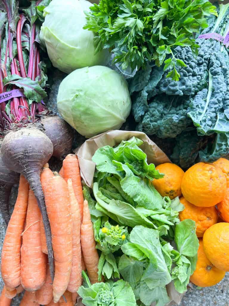Portrait image of late winter produce for Dinner Plans Week 12 featuring red beets, orange carrots, cabbage, mandarins, Napa rabe, and kale.