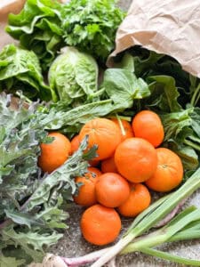 Portrait photo of early spring produce featuring murcott Mandarins, little gems, spinach, kale, and green garlic.