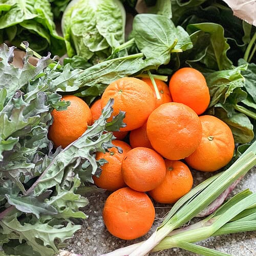 Portrait photo of early spring produce featuring murcott Mandarins, little gems, spinach, kale, and green garlic.